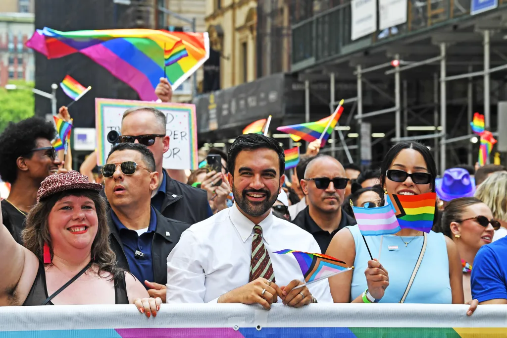 Mayor Zohran Mamdani at a Gay Pride Parade holding a rainbow flag, surrounded by people also holding flags.