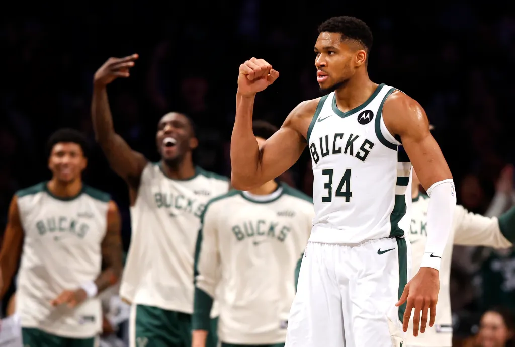 Giannis Antetokounmpo in a white Bucks jersey with number 34, flexing his arm with other players in the background.