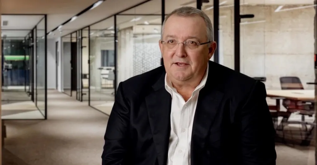 A man in glasses, a white shirt, and a black suit jacket speaking in an office hallway.