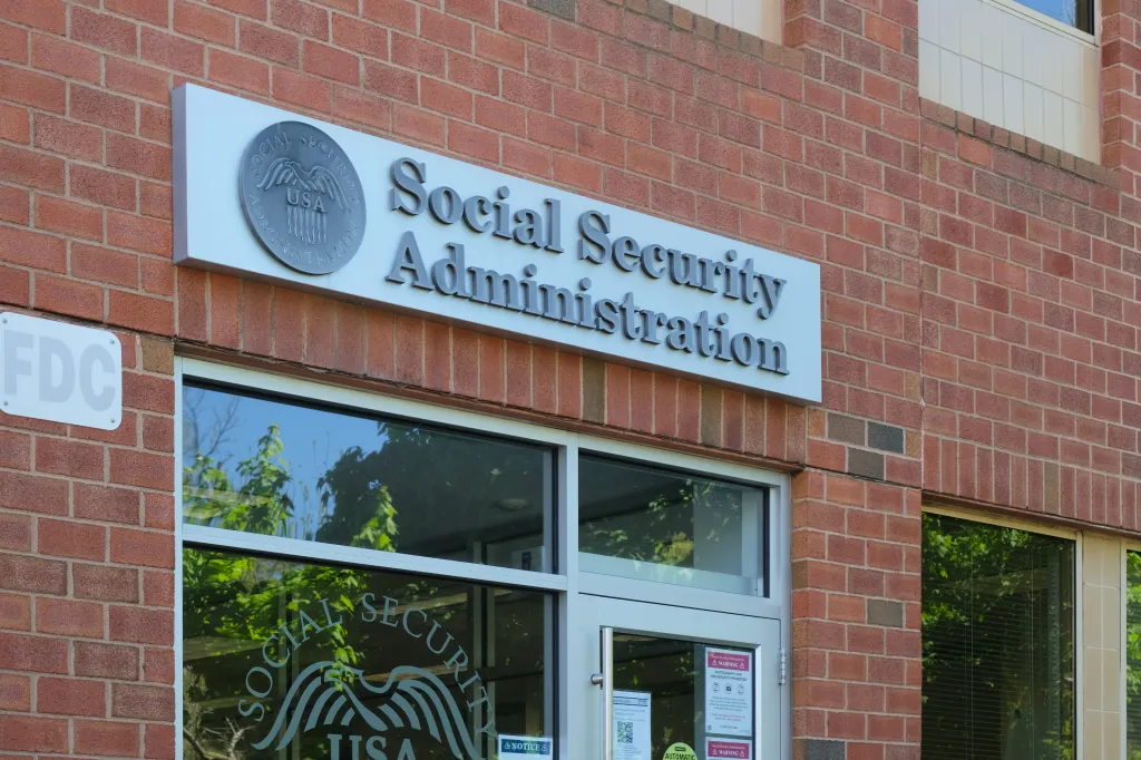 The sign for the Social Security Administration on a brick building, with a reflection of trees in the window.