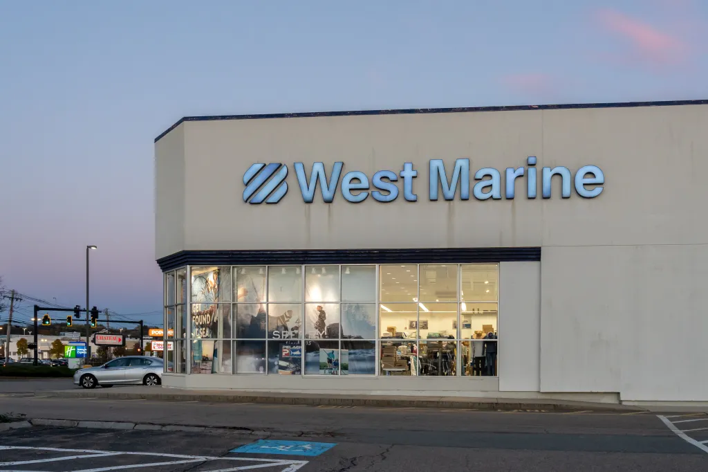 Exterior of a West Marine store in Woburn, MA, with its blue logo above the display windows, at dusk.