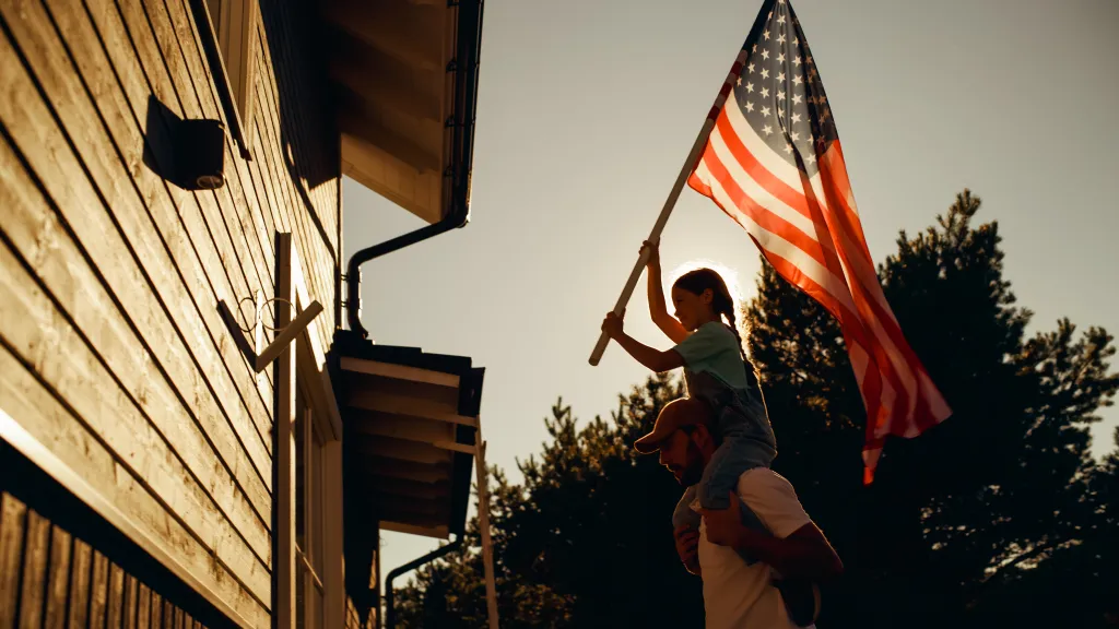 A father carrying his daughter on his shoulders as she holds up an American flag to attach it to their house.