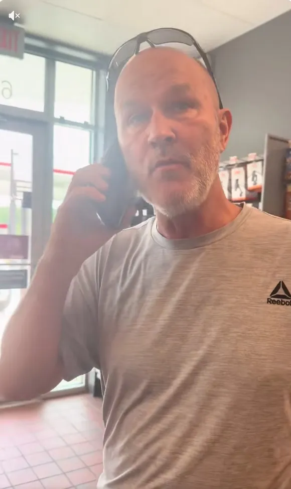 A bald man with a beard wearing a grey Reebok shirt and sunglasses on his head, on the phone, looking forward.