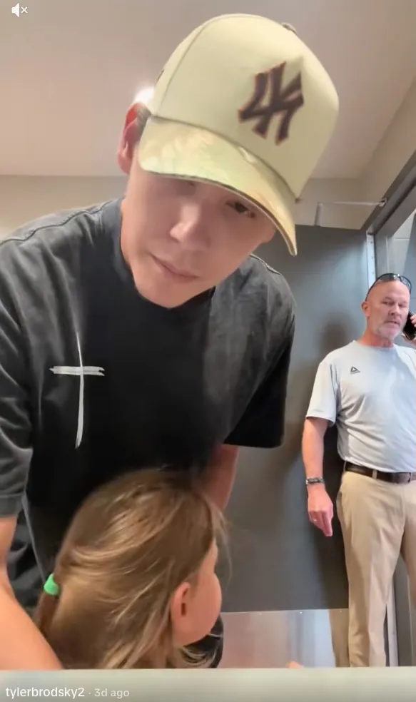 A man wearing a baseball cap with a little girl in front of him, and another man talking on a phone in the background.