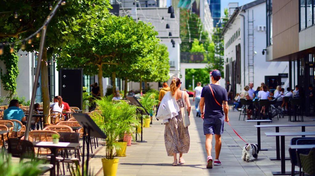 A young couple walking their dog down a city street with outdoor cafes.