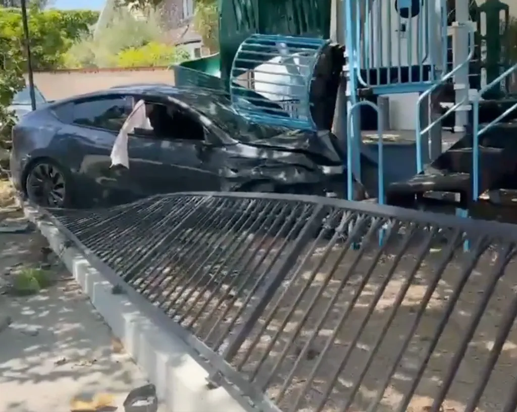 A crashed black Tesla on a playground, with a black metal fence bent in the foreground.