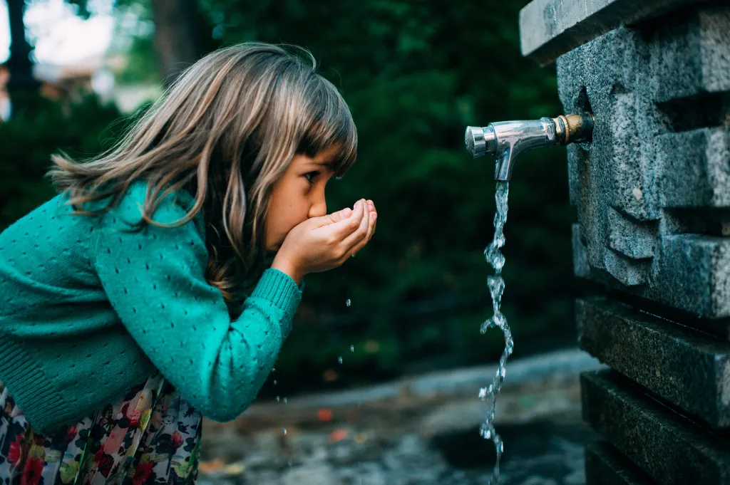A young girl with blonde hair in a teal sweater drinks from a stone water fountain, catching the flowing water in her cupped hands.
