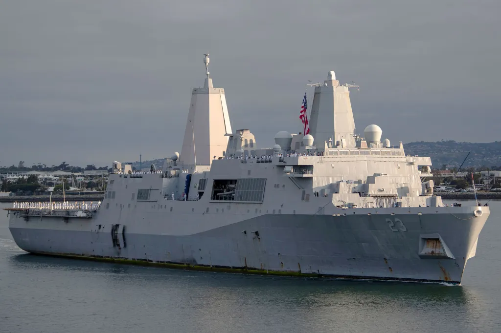 USS Anchorage (LPD-23) in the water, with sailors lining its deck and an American flag flying from its mast.