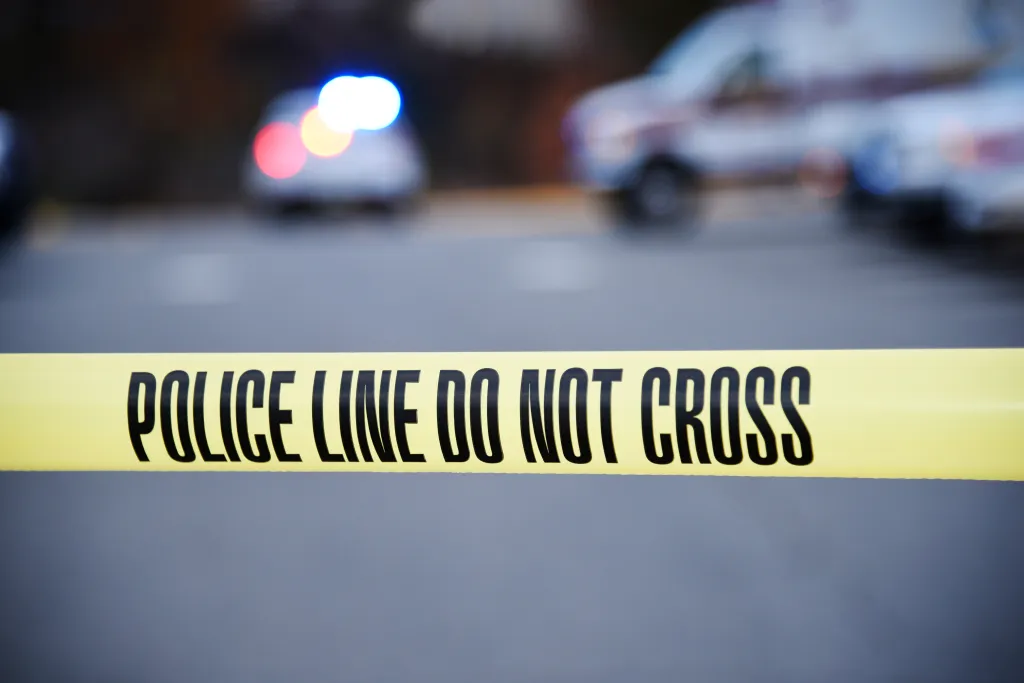 Yellow police tape with "POLICE LINE DO NOT CROSS" in black text, with blurred police cars in the background.