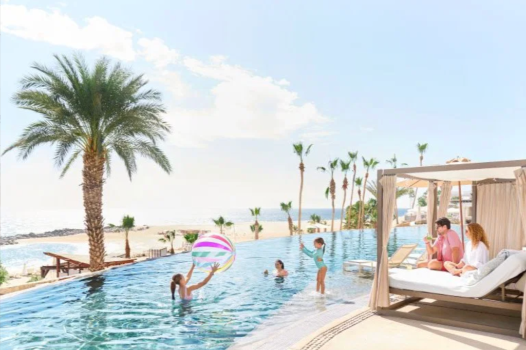 Families enjoying a Hilton hotel stay, with adults relaxing in a cabana next to an infinity pool while children play with a beach ball in the water.