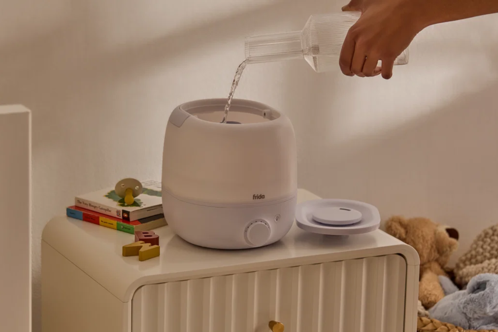A hand pouring water into a Frida Baby Cool Mist Humidifier on a nightstand next to children's books and a pacifier.