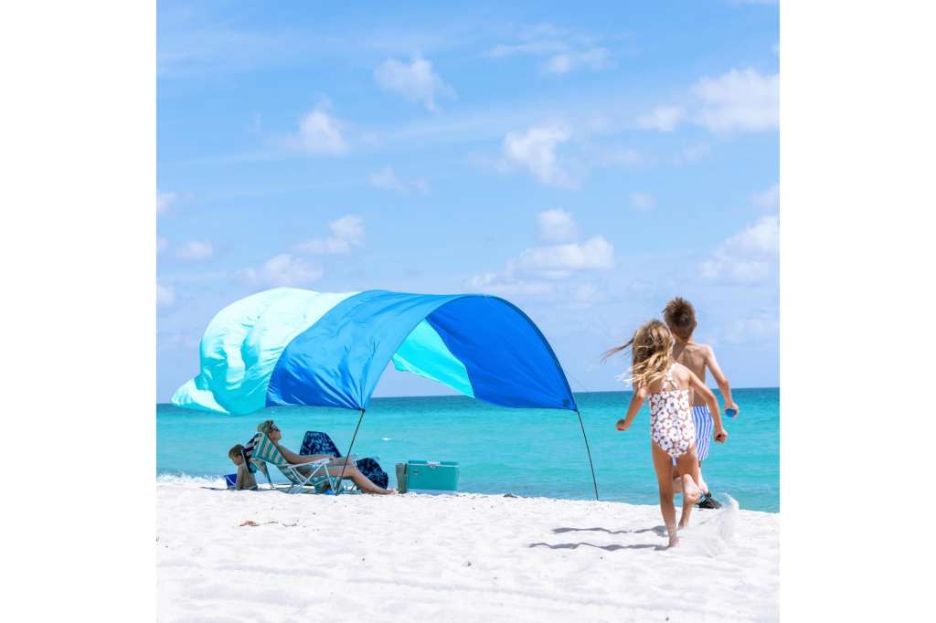 A blue and turquoise Shibumi Shade canopy provides shade for a woman and child sitting on a white sand beach, while two children run towards the ocean.