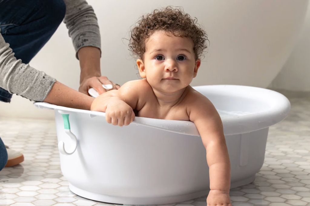 A baby with curly hair looks at the camera while being bathed in a white Frida Baby Tub.