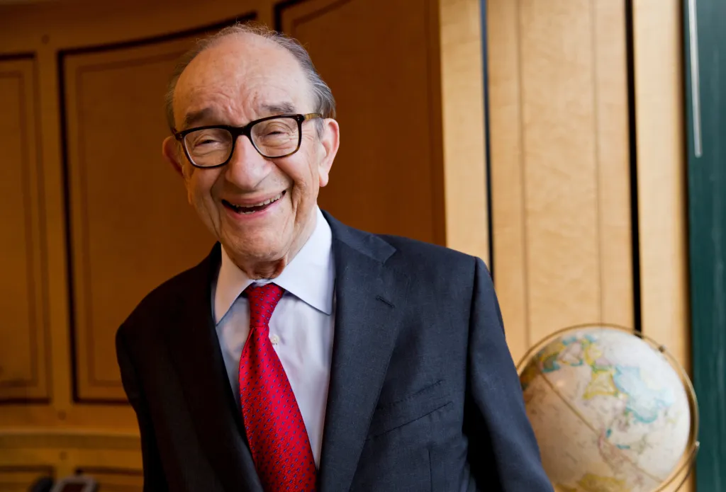 Economist Alan Greenspan smiles, wearing glasses, a suit jacket, and a red tie.