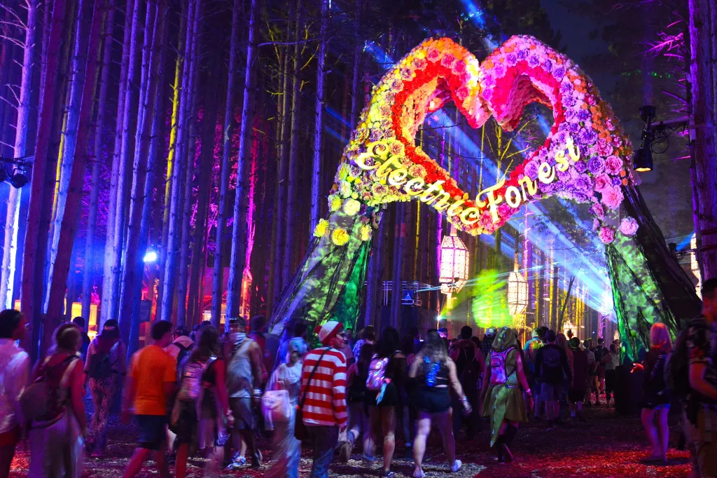 Attendees walk through a forest illuminated with colorful lights and a heart-shaped archway at the Electric Forest Festival.