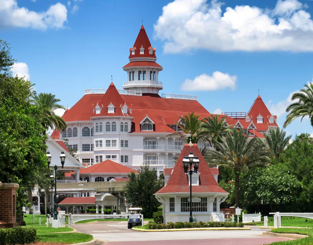 A view of Disney's Grand Floridian Resort main entrance, with a white, Victorian-style building featuring a red roof and several towers, surrounded by palm trees and lush greenery.