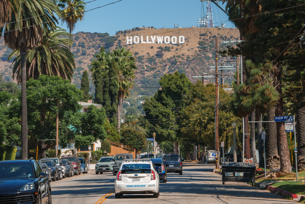 The Hollywood Sign is visible behind palm trees and residential buildings on a sunny day in Los Angeles.