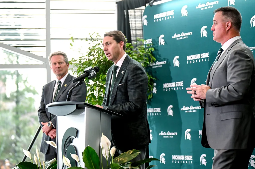 MSU Athletic Director J Batt speaking at a podium with two other men, one on each side.