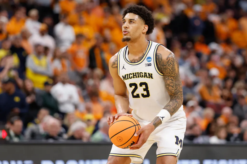 A male Michigan Wolverines basketball player in a white jersey and shorts with number 23, holding a basketball.