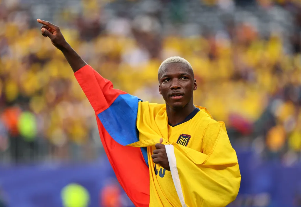 Moisés Caicedo with an Ecuador flag draped over his shoulders, raising his left arm and pointing.