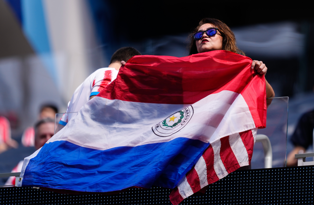 A fan wearing sunglasses holds up a Paraguayan flag at a soccer match.