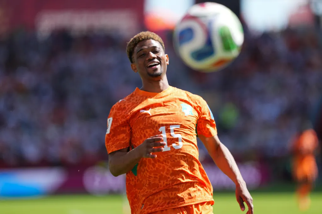 Amad Diallo of Ivory Coast, wearing an orange jersey with "15" on it, smiling as he looks up at a soccer ball in the air.