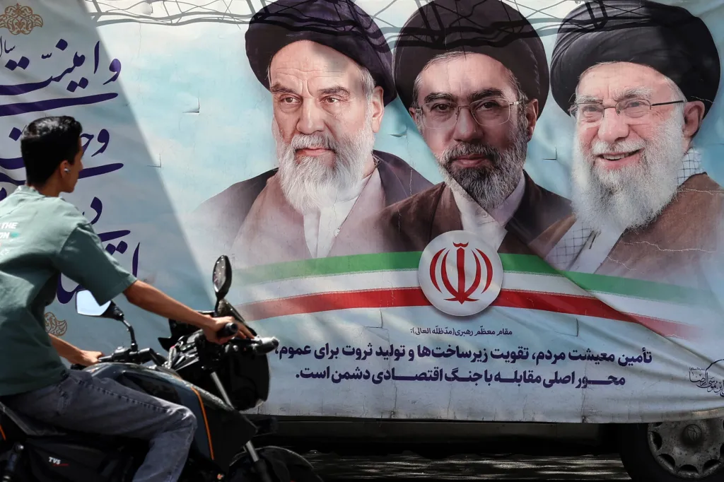 An Iranian man on a motorbike rides past a billboard with images of Ruhollah Khomeini, Mojtaba Khamenei, and Ali Khamenei, along with the Iranian flag and Farsi text.
