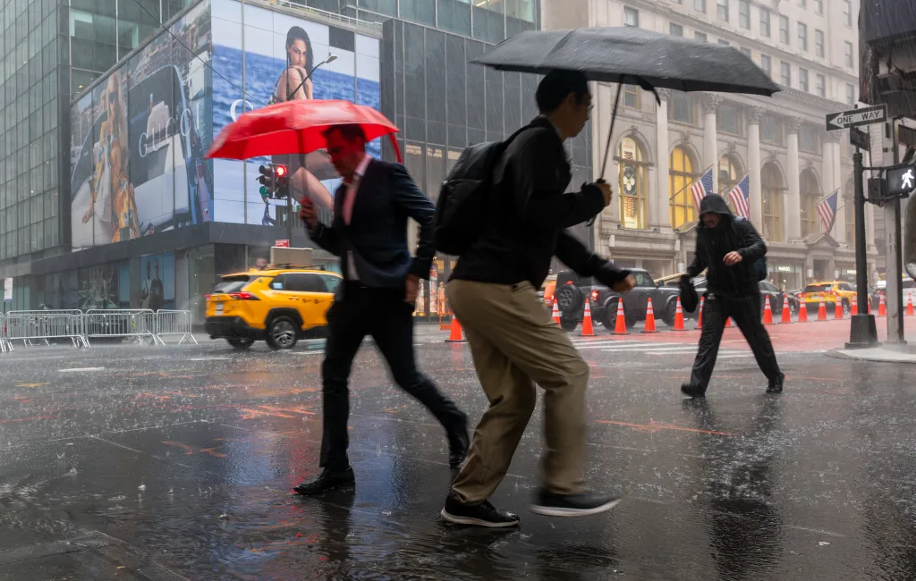 People walking through a rainy New York City street with umbrellas, with a yellow taxi cab and buildings in the background.