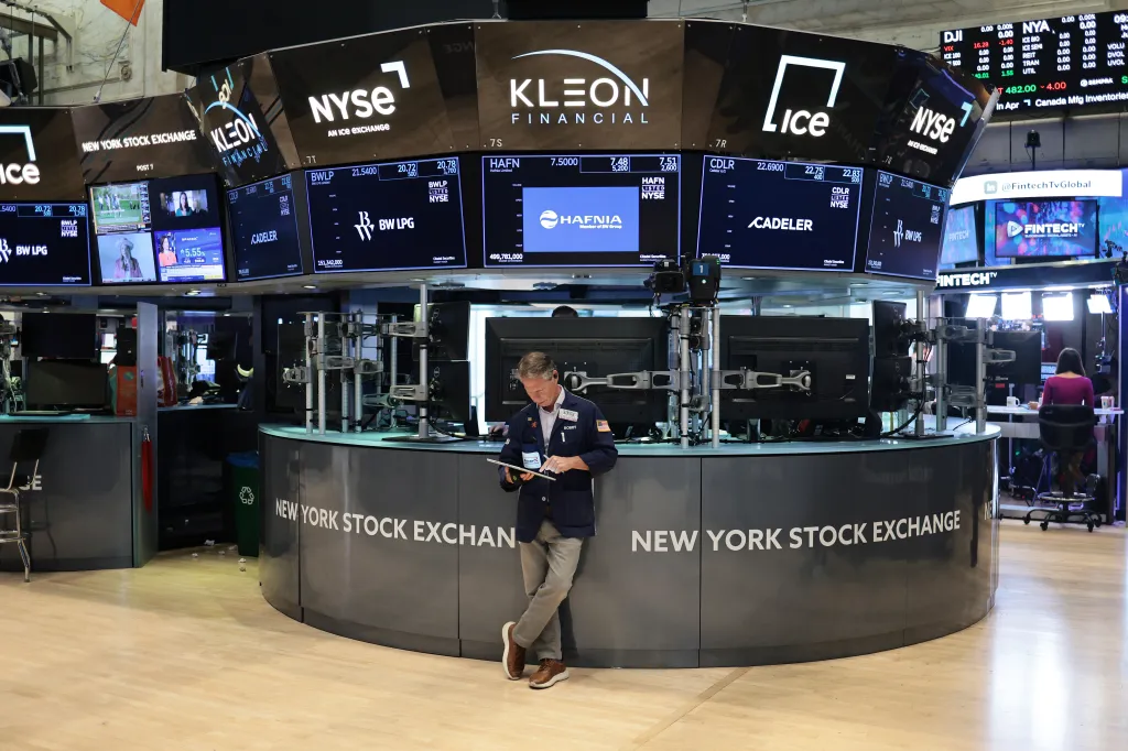 A trader looking at a tablet in front of large screens displaying stock prices at the New York Stock Exchange.