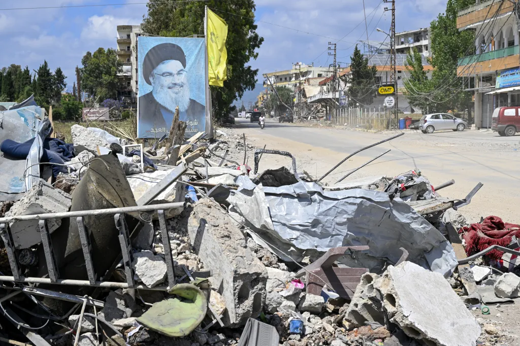 Debris, including concrete, metal, and wood, litters the foreground, with a large poster of a bearded man in a turban and a yellow flag behind it. In the background, damaged buildings line a road where vehicles are present, under a cloudy sky.