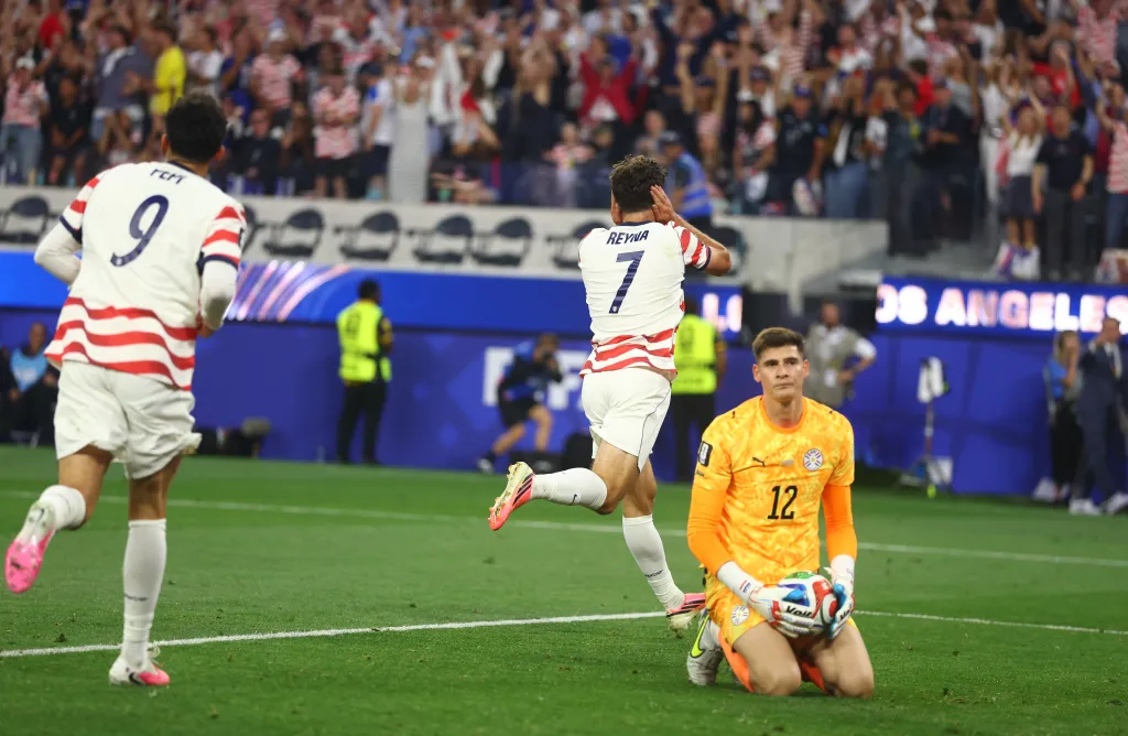 Soccer players celebrate while a goalkeeper kneels on the field.