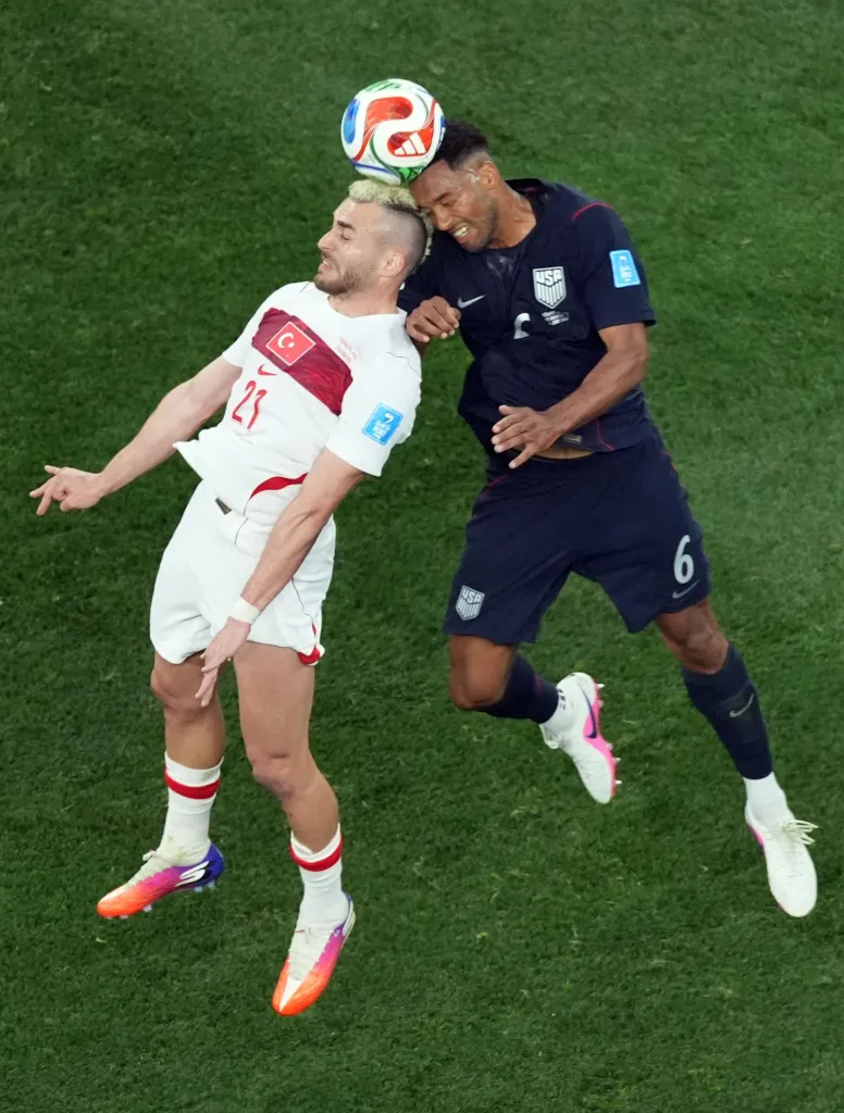 Two soccer players, one from Turkey and one from the USA, jump for a header.