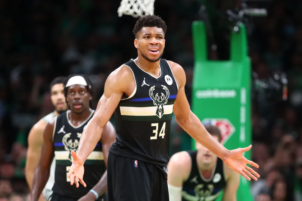 Giannis Antetokounmpo in a black Milwaukee Bucks jersey, arms outstretched, during an NBA game.