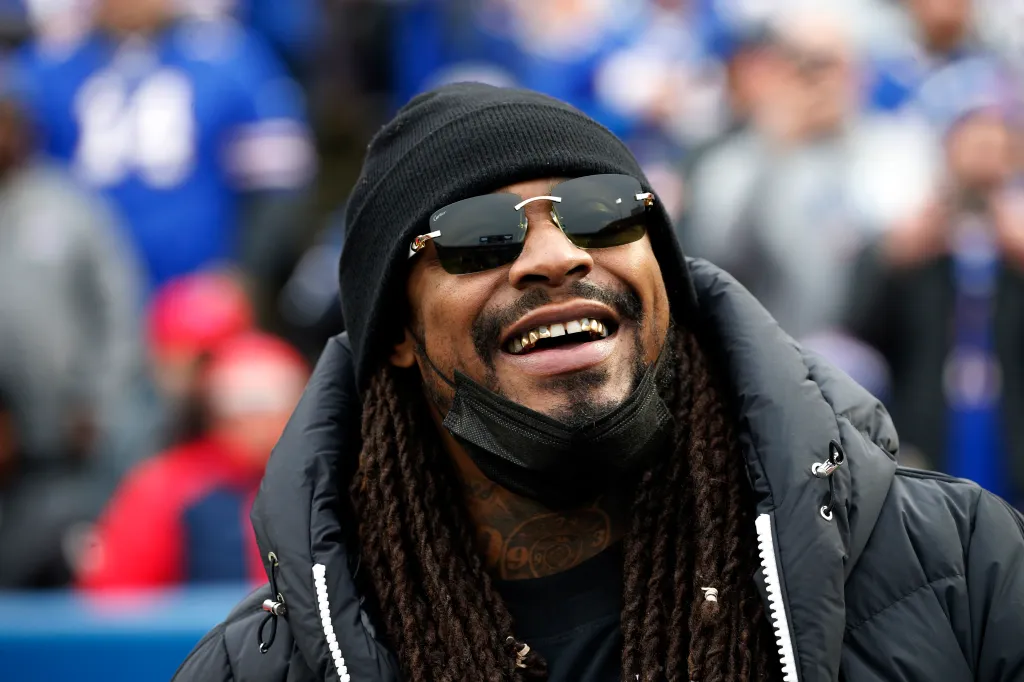 Marshawn Lynch smiling and wearing a black beanie and sunglasses.