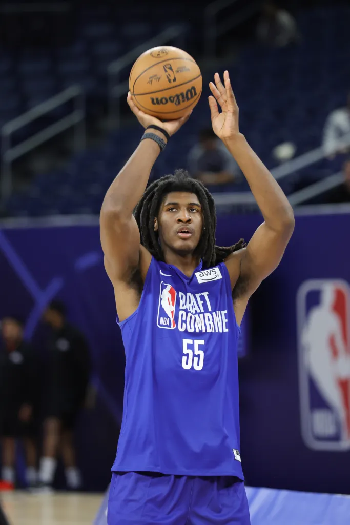 A basketball player shoots a basketball while wearing a blue jersey with "Draft Combine 55" written on it.