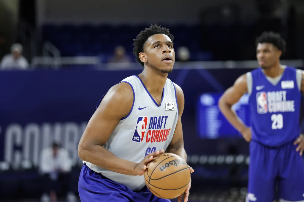 A basketball player, wearing a white jersey with "NBA Draft Combine" on it, holds a basketball and looks up, with another player in a blue jersey in the background.