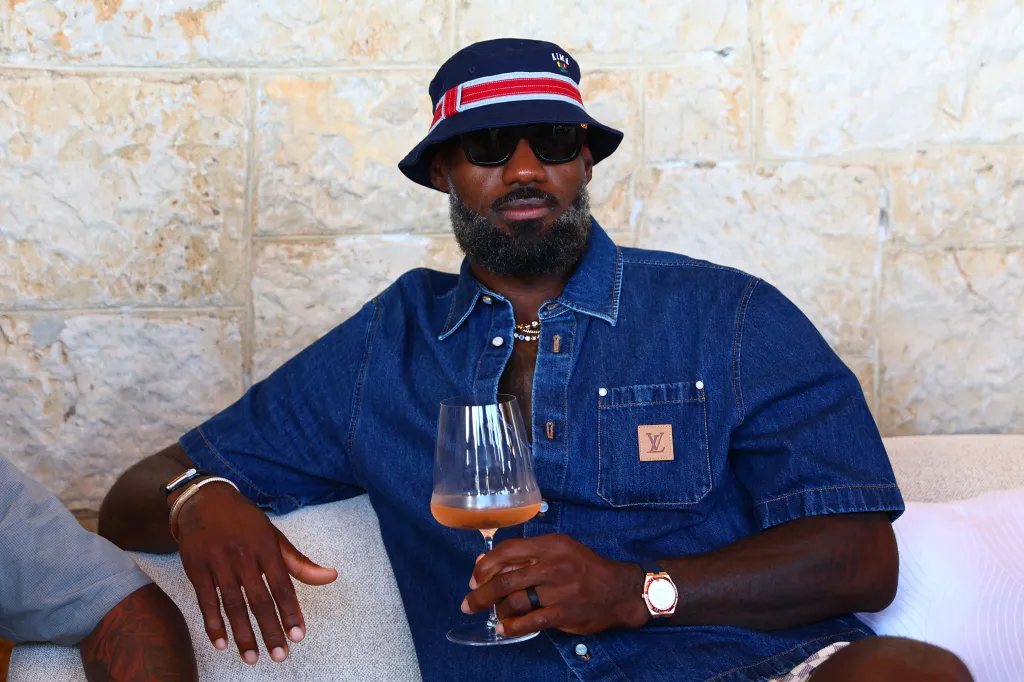 Man in a bucket hat, sunglasses, and denim shirt holding a glass of rose wine.