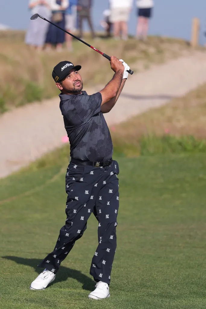 A male golfer in a gray shirt and patterned pants swings a club with his back to the viewer.