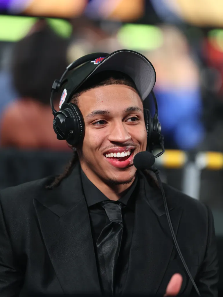 A man wearing a black suit, cap, and headphones with a microphone, smiling and looking to the right.
