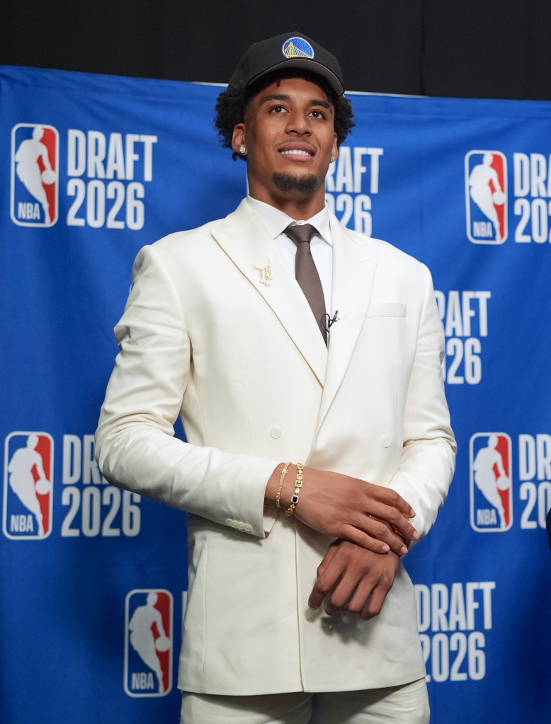 A young man in a cream suit and a Golden State Warriors cap stands smiling in front of an NBA Draft 2026 backdrop.
