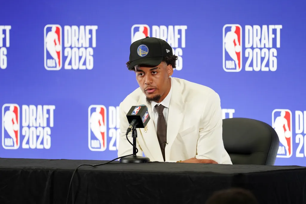 A draftee wearing a Golden State Warriors cap and a white suit, sitting in front of a blue backdrop with "DRAFT 2026" repeated.