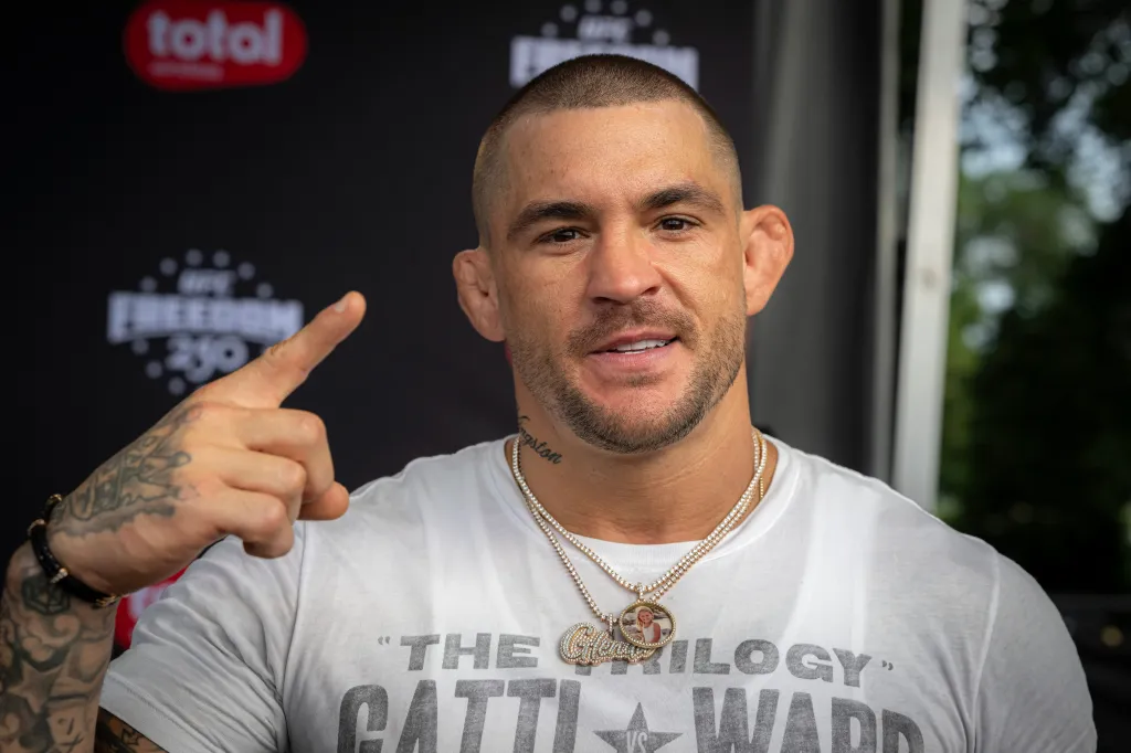 UFC fighter Dustin Poirier with his right arm bent, index finger pointed up, wearing a white t-shirt and two gold chain necklaces.