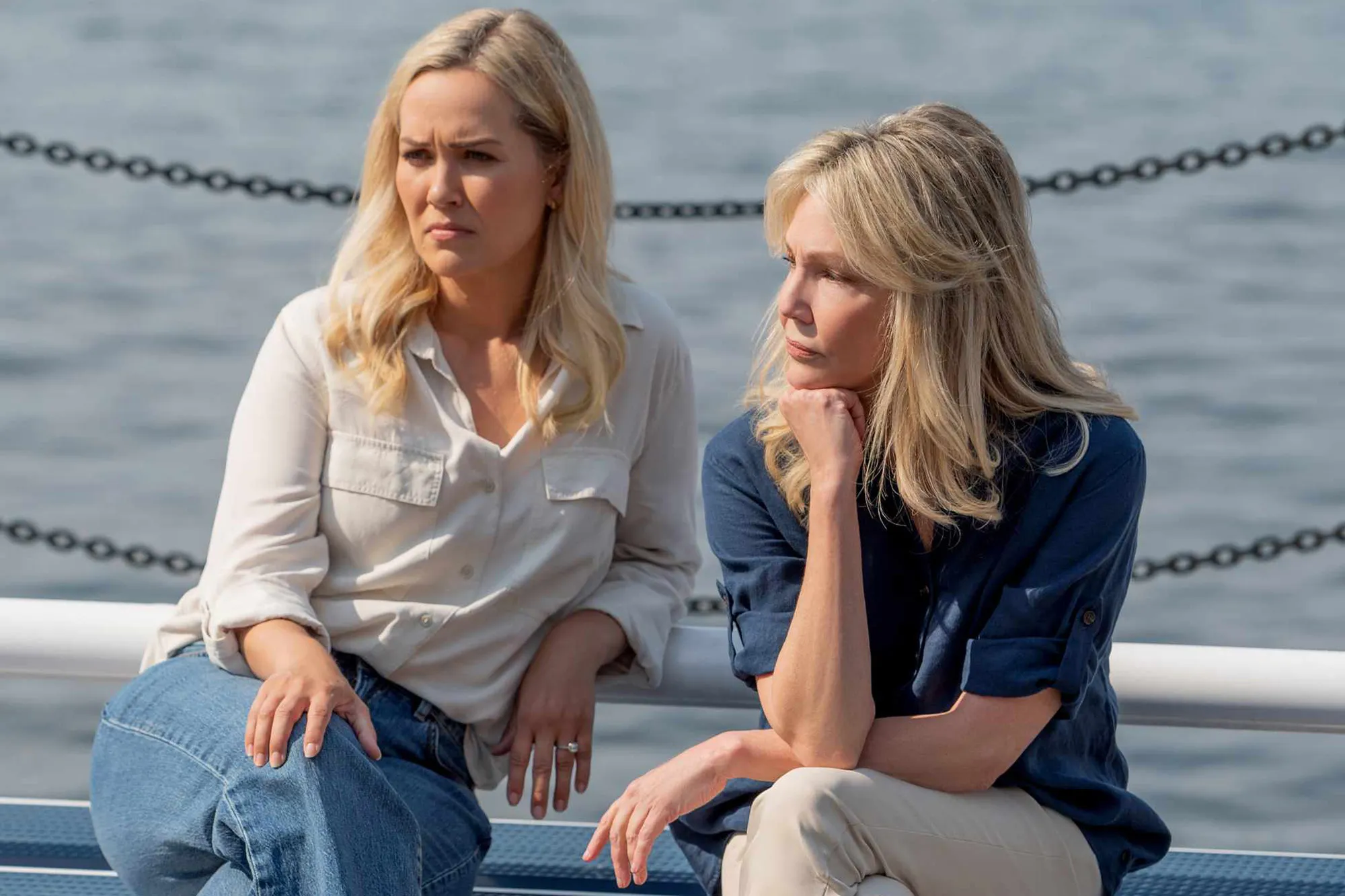 Two women, one in a light shirt and jeans and the other in a blue shirt and khaki pants, sit looking contemplative by the water.