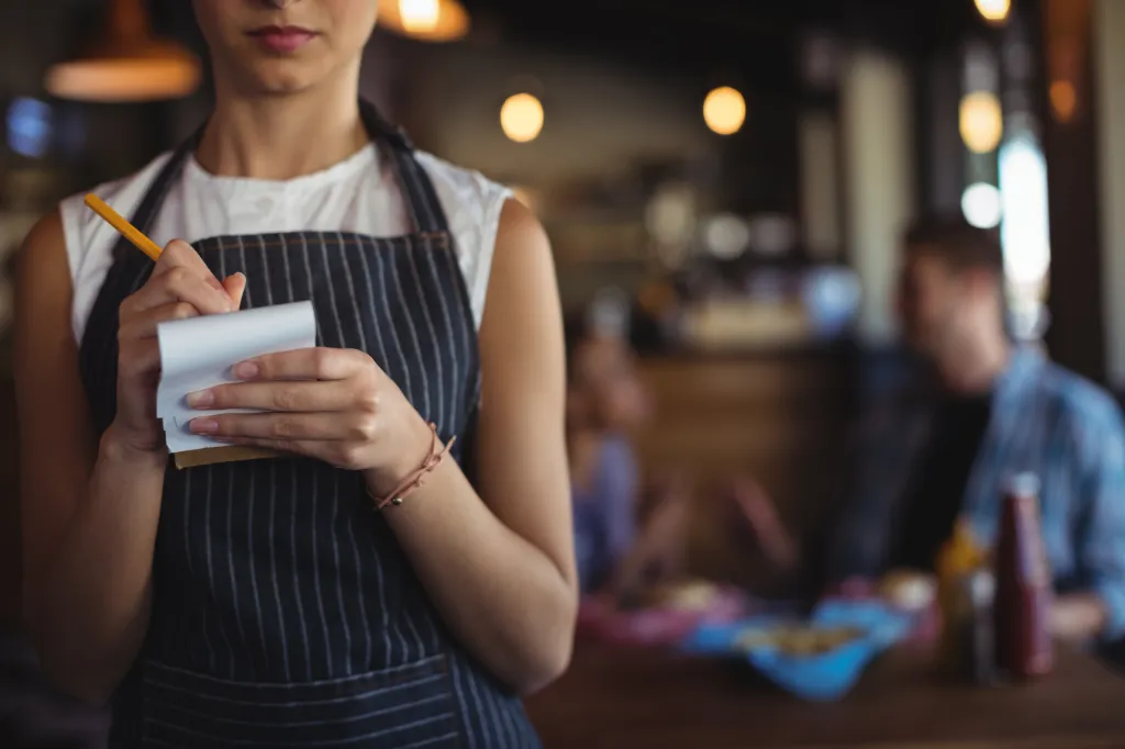 A waitress in a striped apron takes a customer's order on a notepad with a pencil.