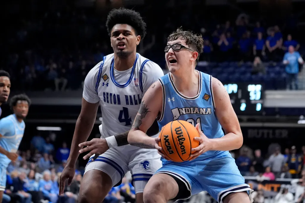 A basketball player in a light blue jersey with "INDIANA" written on it and wearing goggles, holding a basketball while being guarded by a player in a white Seton Hall jersey.