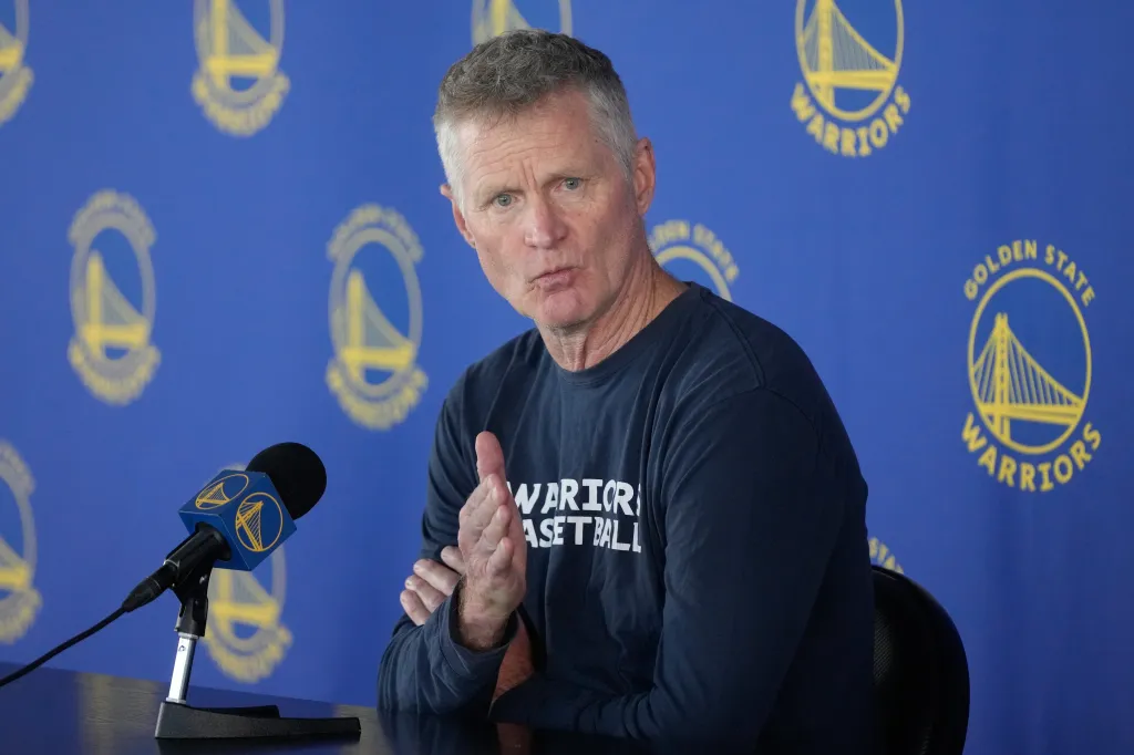 Steve Kerr speaking at a press conference with a microphone in front of a blue backdrop with Golden State Warriors logos.