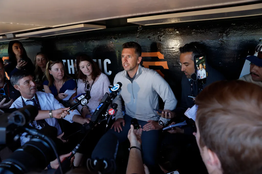 A man in a light grey shirt surrounded by reporters holding microphones.