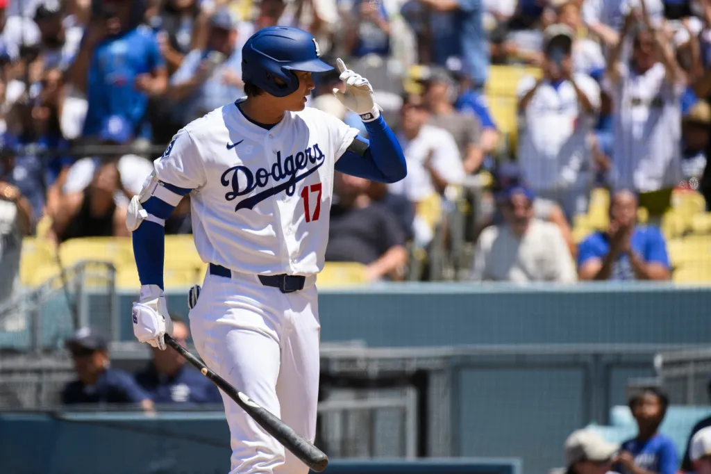 Shohei Ohtani in a Dodgers uniform with a batting helmet and bat, looking to the side.