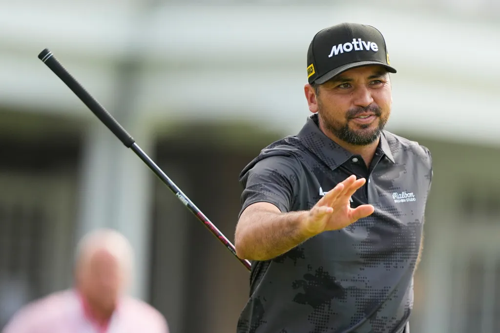 Jason Day wearing a black cap with a white "motive" logo and a gray polo shirt, holding a golf club and gesturing with his right hand.