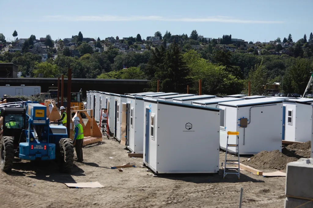 Temporary shelters for the homeless being constructed, with workers and equipment on a dirt lot.